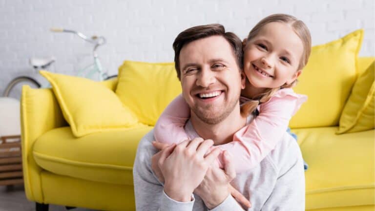 Adult man laughing while cheerful daughter hugging him at home
