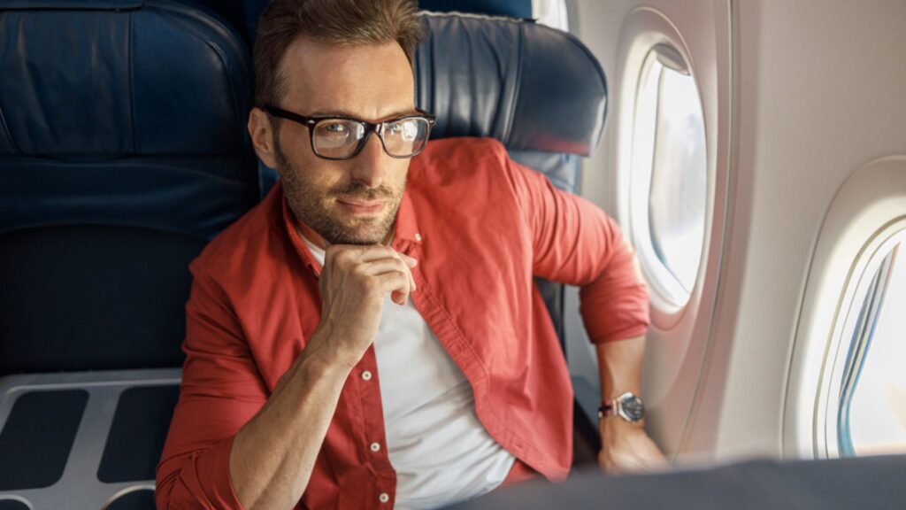Attractive caucasian businessman in casual wear and glasses looking thoughtful, sitting on the plane near the window