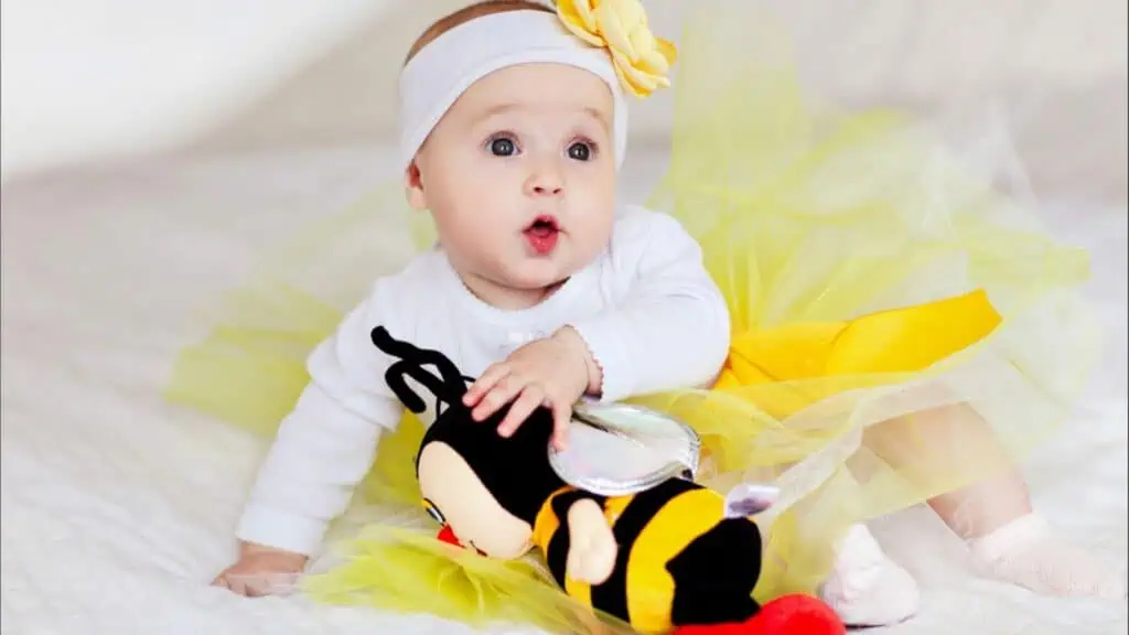Baby in a yellow skirt sits on the bed with a toy bee