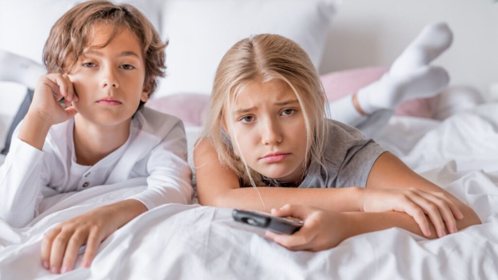 Boring boy and girl in pajamas lying in bedroom at home watching tv feeling bored