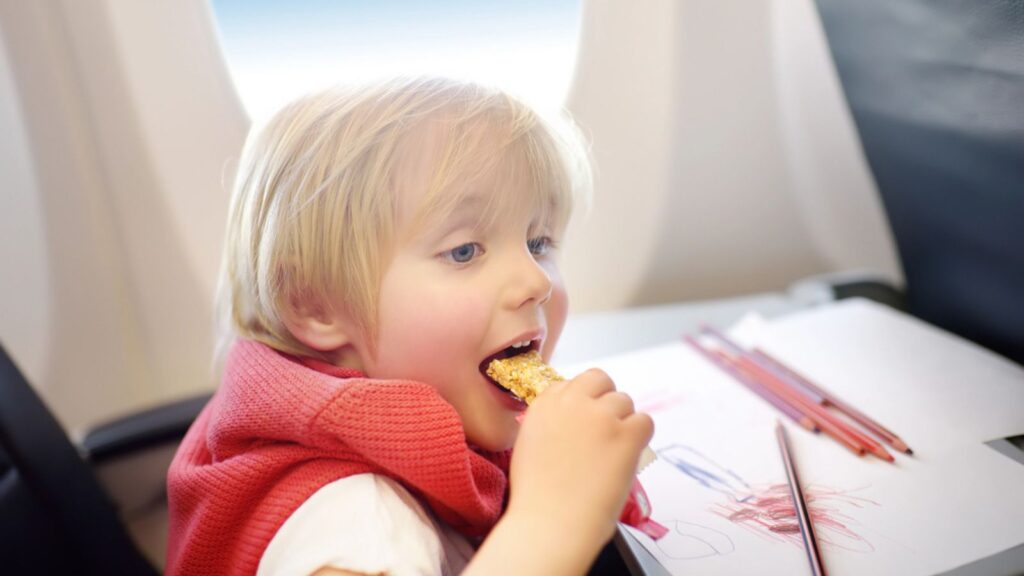 Charming kid traveling by an airplane. Joyful little boy sitting by aircraft window during the flight. Child drawing picture and eating snack
