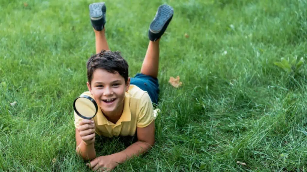 Cheerful boy lying on grass and holding magnifier