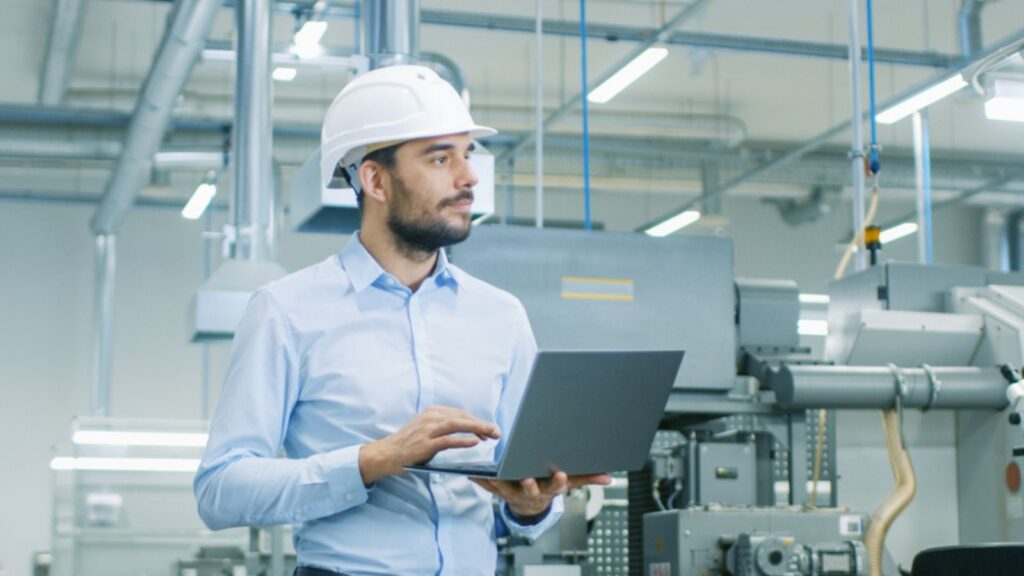 Chief Engineer in the Hard Hat Walks Through Light Modern Factory While holding laptop