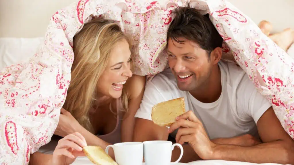 Couple Snuggled Under Duvet Eating Breakfast