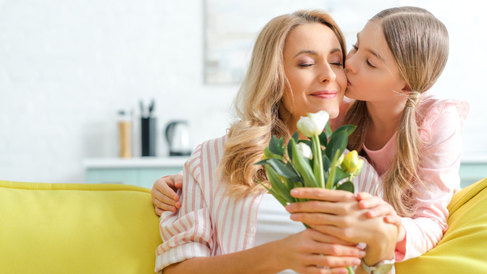 Cute daughter kissing happy mature mother holding bouquet of tulips