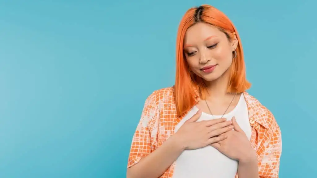 Grateful young asian woman with dyed red hair smiling and holding hands over her heart