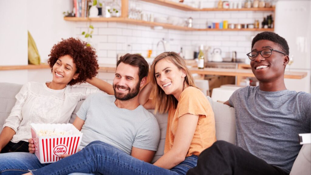 Group Of Friends Sitting On Sofa And Watching Movie At Home While eating popcorn