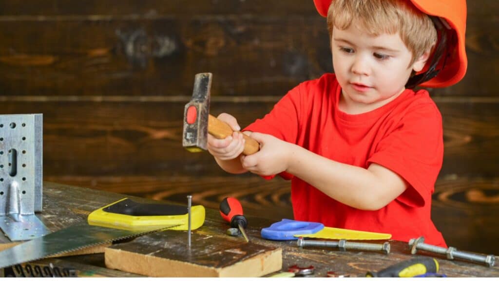 Handcrafting concept. Toddler boy on busy face plays with hammer tool at home in workshop