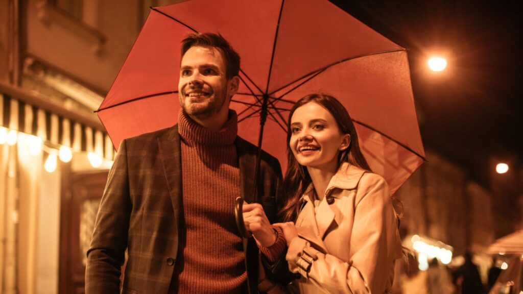 Happy couple in autumn outfit walking under umbrella along evening street