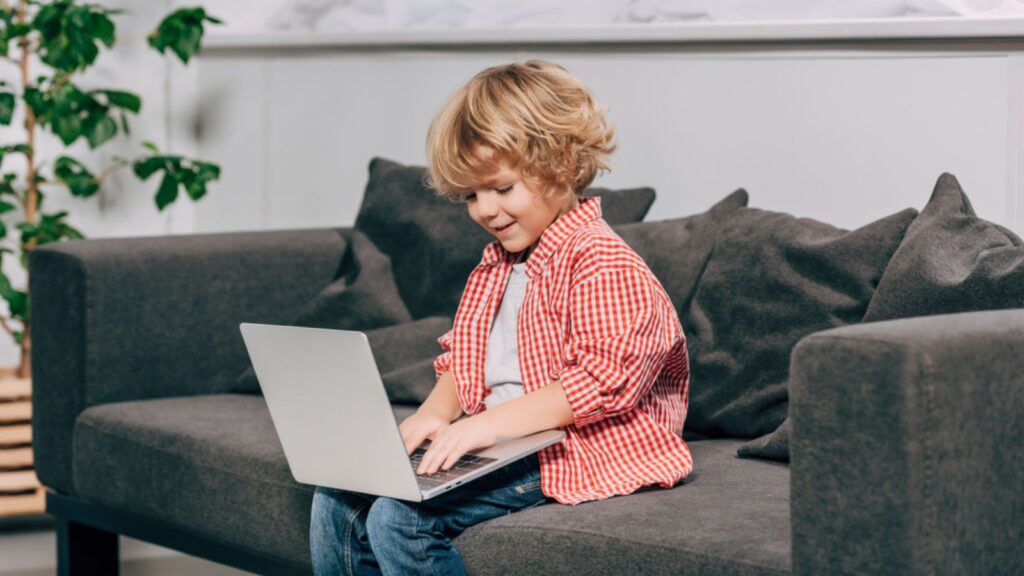 Happy curly boy using laptop on sofa at home