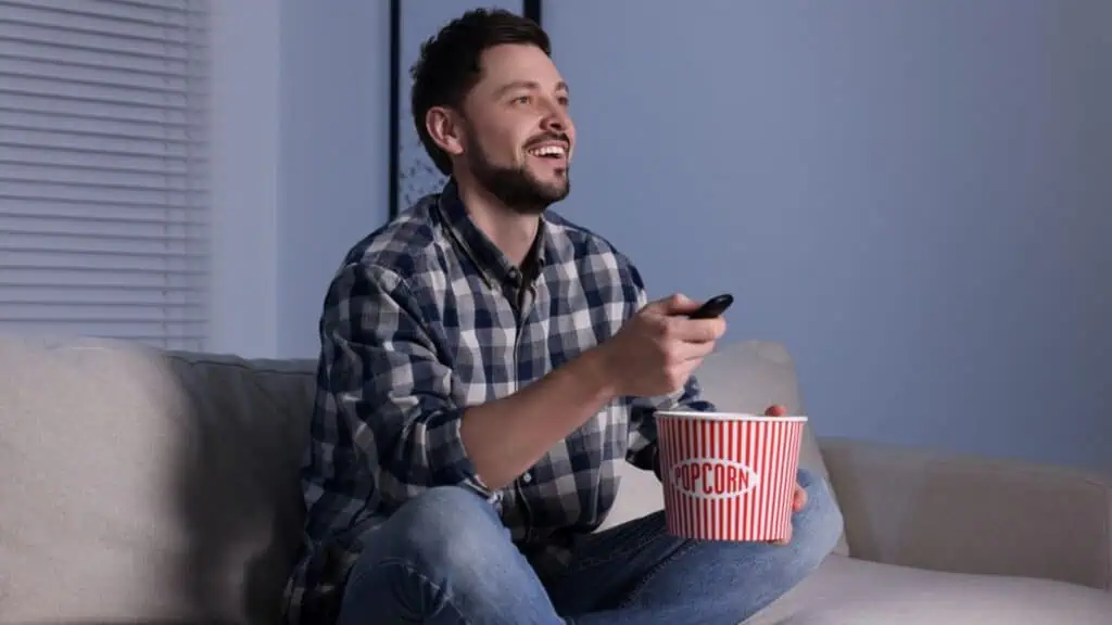 Happy man with popcorn bucket changing TV channels with remote control at home in evening