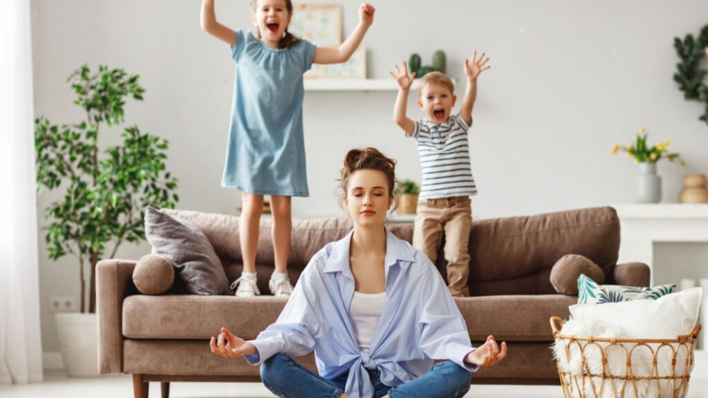 Happy mother with closed eyes meditating in lotus pose on the floor trying to be calm to her kids