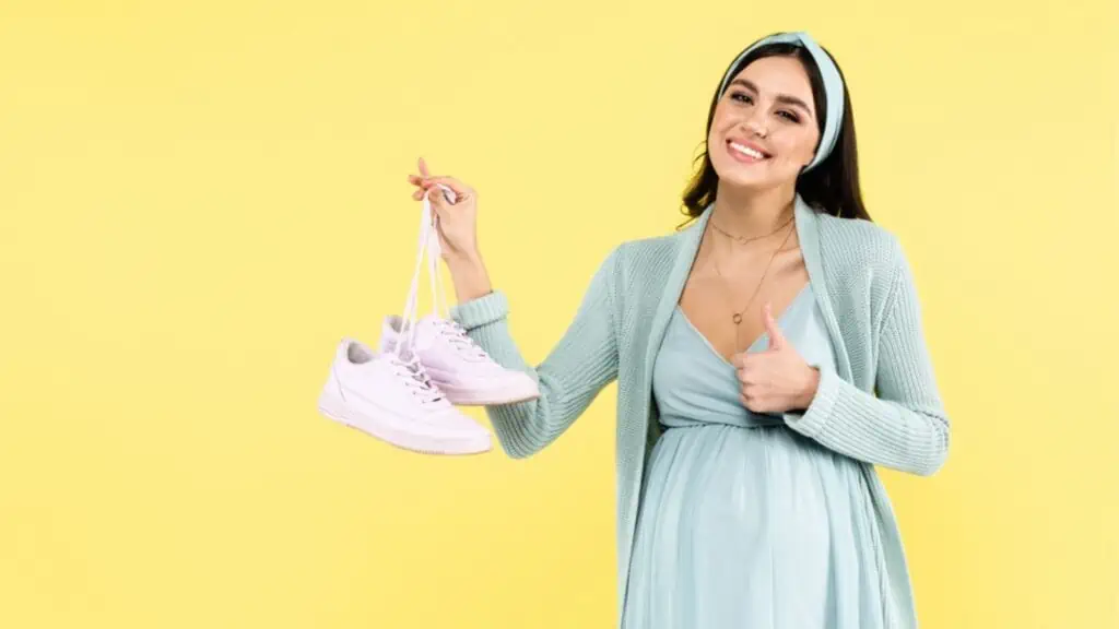 Happy pregnant woman showing thumb up while holding sneakers isolated on yellow
