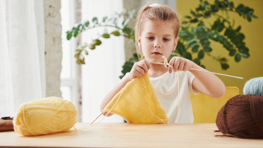 Kid is knitting at home. Cute little girl sitting near the wooden table is learning some new stuff