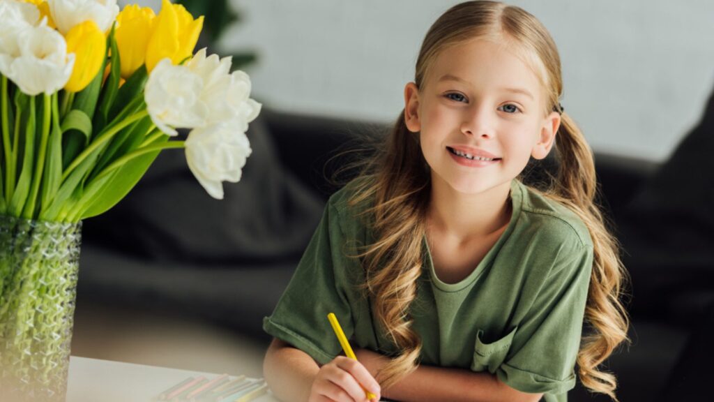 Little girl drawing in a paper with a flower vase in the table