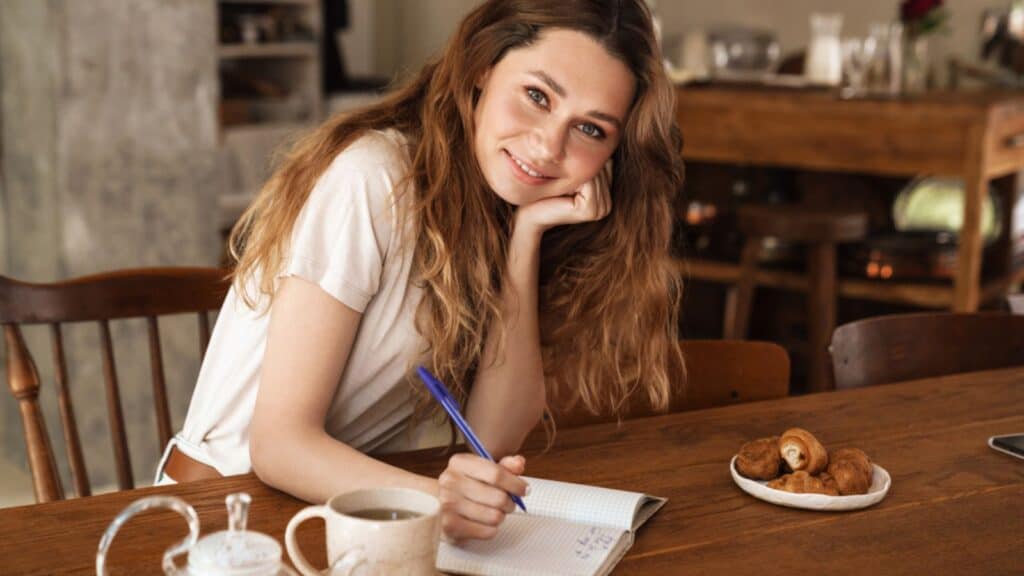 Lovely cheerful woman sitting at the kitchen table writing notes in her diary