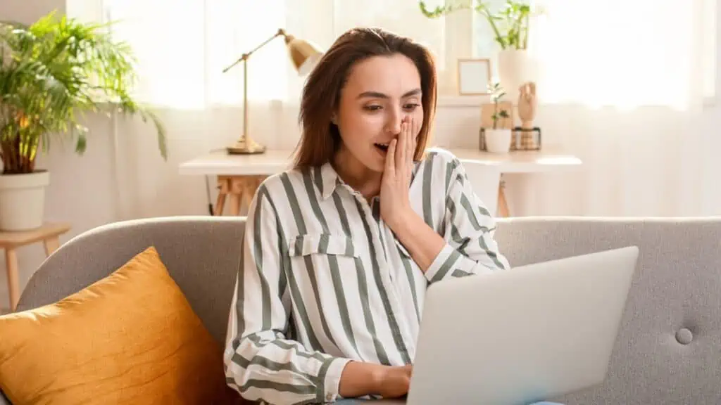 Shocked woman with laptop reading e-mail message on sofa in light room