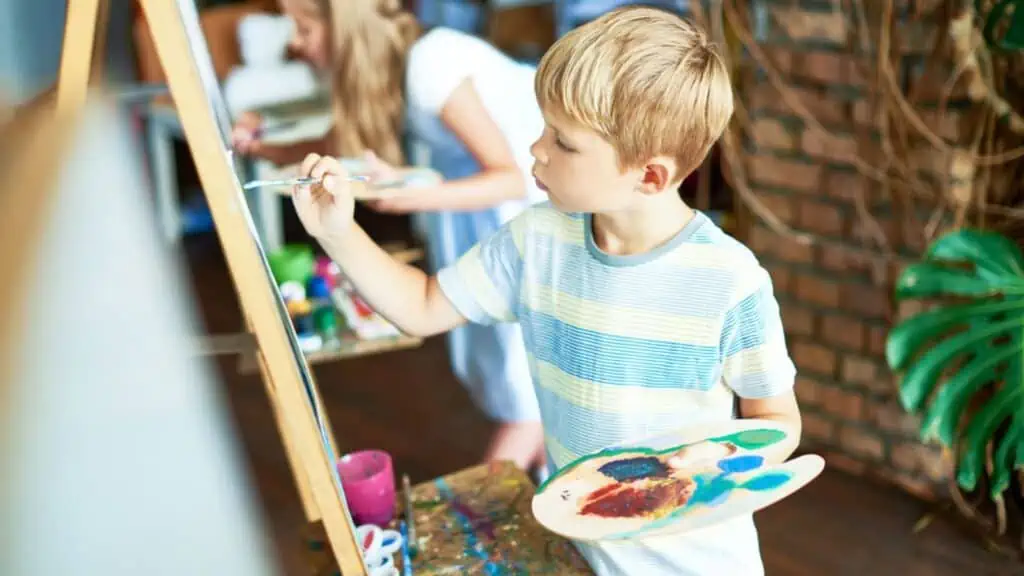 Side view portrait of blonde little boy painting on easel enjoying art class
