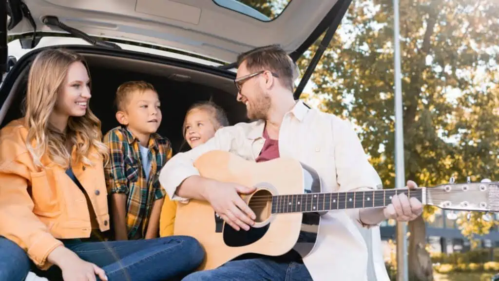 Smiling father playing acoustic guitar near wife and kids in car