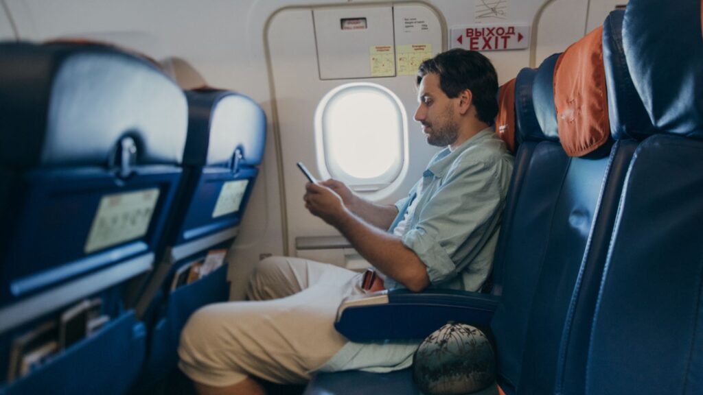 The guy is flying in an airplane with a phone in hand