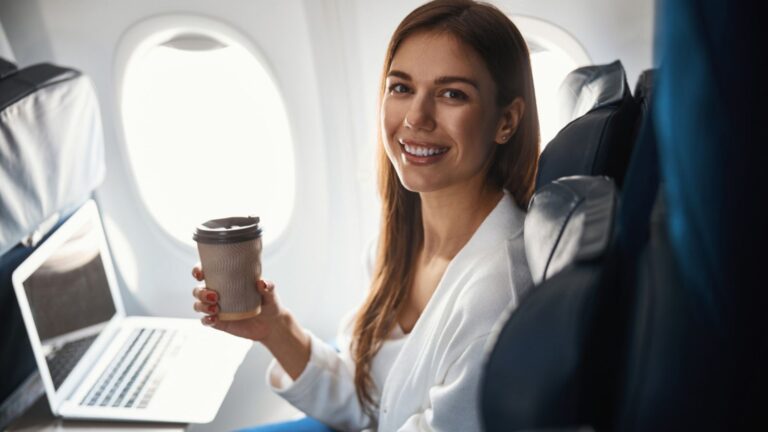 Woman seated at the window side in airplane drinking hot beverage with laptop