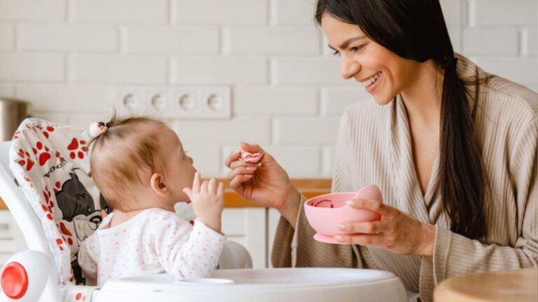 Young brunette mother smiling and feeding her baby in kitchen at home