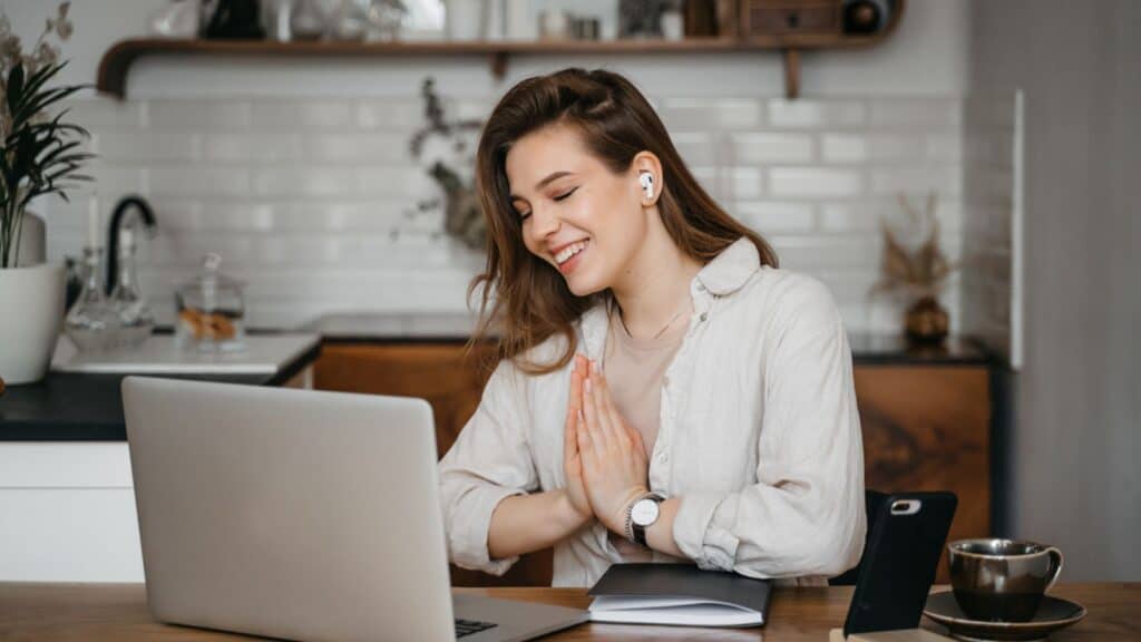 Young pretty woman talking on video call and waving hand while sitting at table in the kitchen